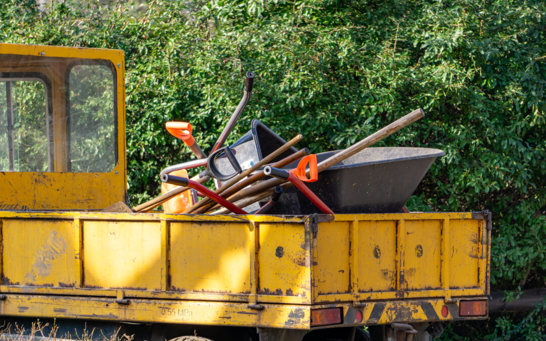 A small landscaping truck with gardening tools piled up in the back such as shovels, rakes, and a wheelbarrow