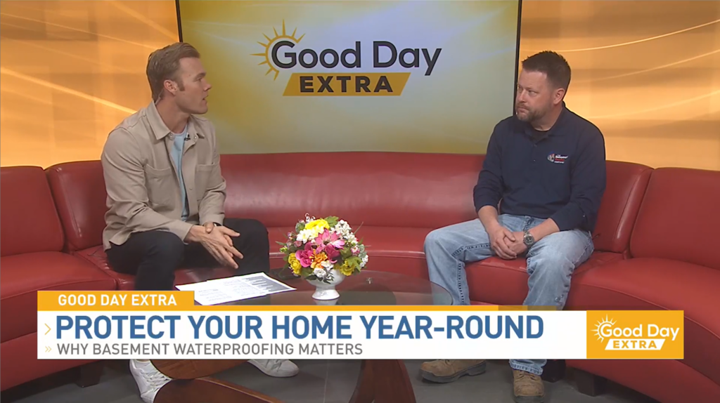 Two people sit on a red couch during a “Good Day Extra” TV segment discussing basement waterproofing, with on‑screen text reading “Protect Your Home Year‑Round.”